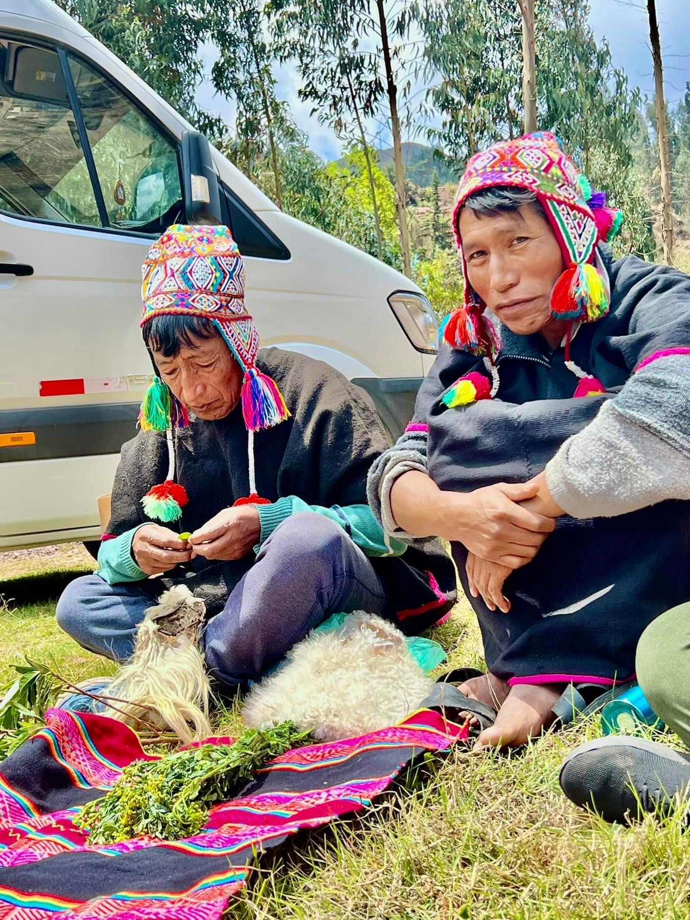 Two people seated beside a woven cloth and ceremonial objects on grass