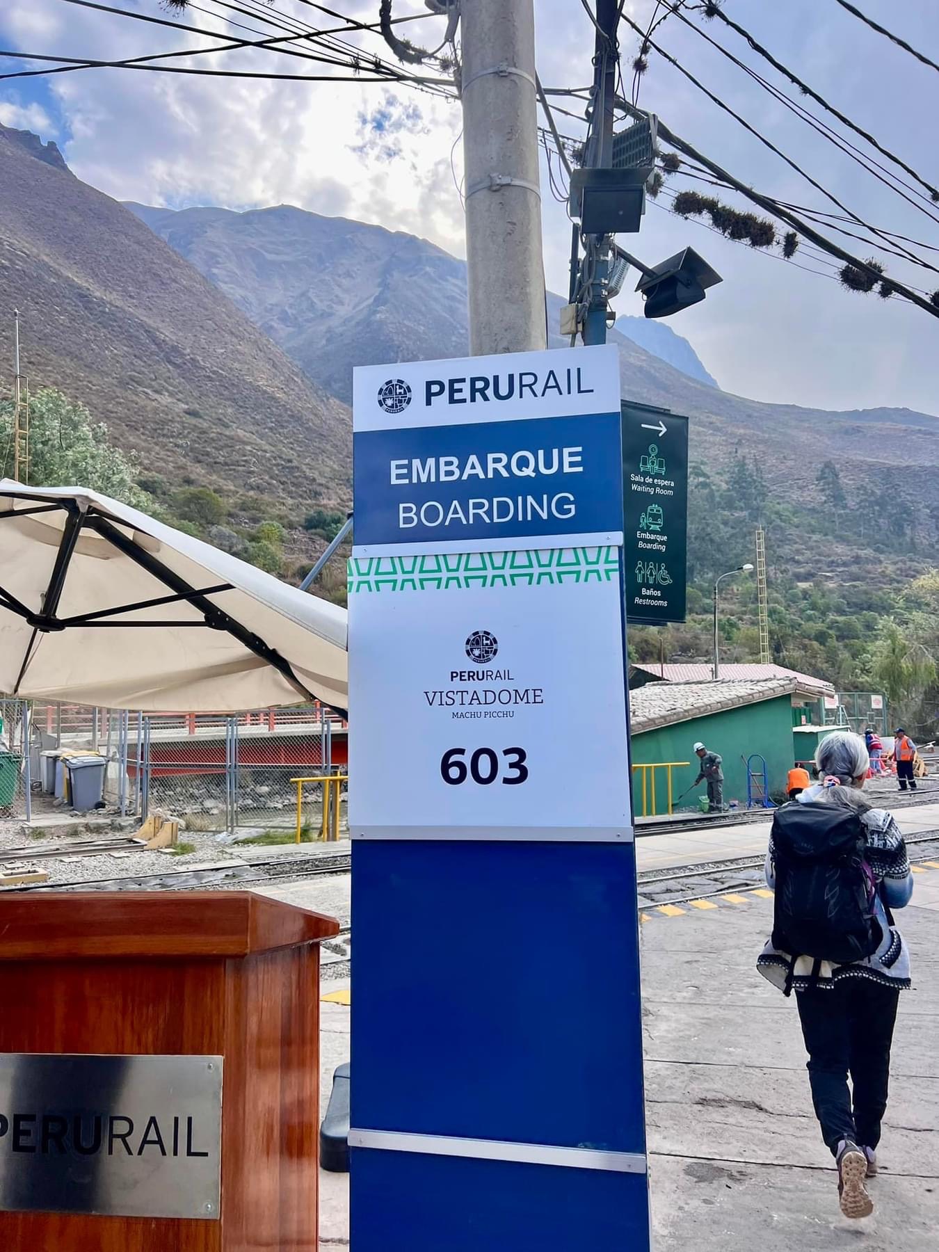 Armanda standing beneath an embarkation sign at a train platform with mountains behind her