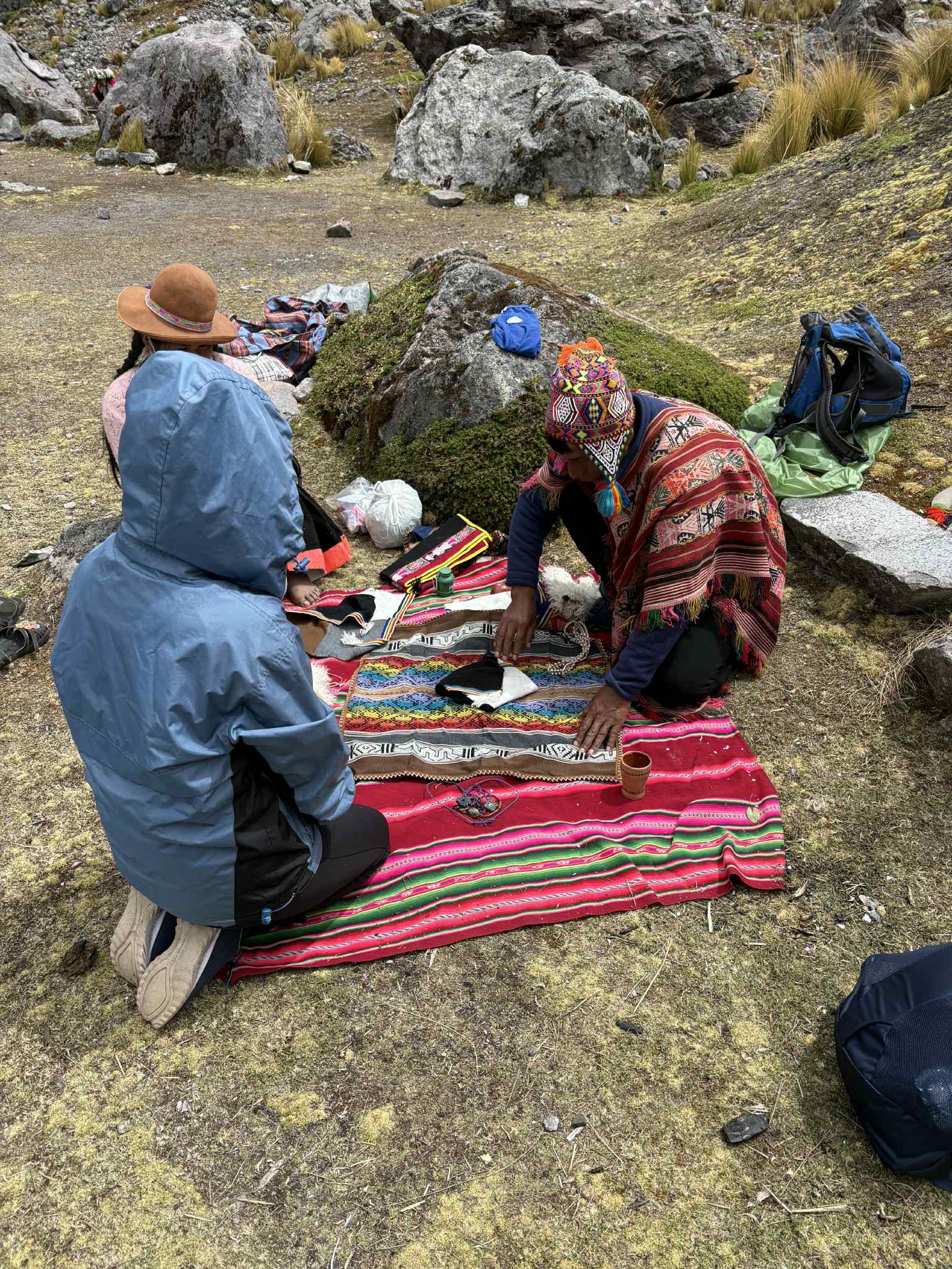 People seated around a striped woven cloth and ceremonial objects in a rocky mountain clearing